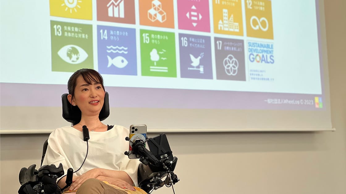 代表・織田友理子が講演でSDGsの目標を示すスクリーンの前で話す様子