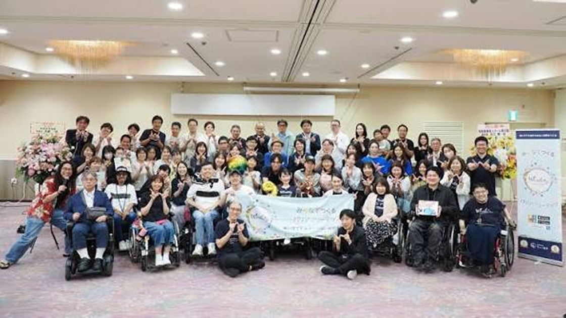 室内で開催されたイベントで多くの参加者と車いすユーザーが集合写真に写る様子